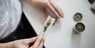 Person die einen Joint dreht vor einem weißen Tisch auf dem ein Grinder steht