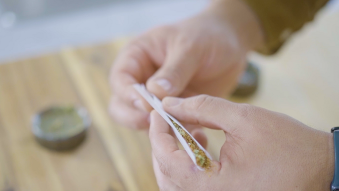 Zwei Hände die einen Joint zusammendrehen, im Hintergrund unscharf ein Holzbrett mit einem Grinder