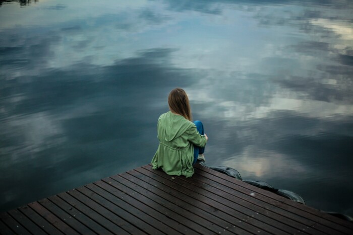 Frau mit Jacke sitzt am See und schaut auf die Wasseroberfläche