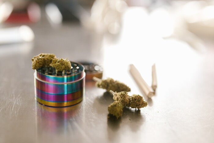 Bunter Grinder mit Cannabisblüten welcher neben zwei Joints auf einem hellen Tisch liegt