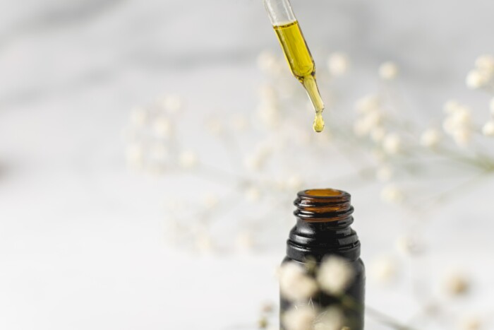 Eine mit Öl gefüllte Pipette welche aus einem Behälter Öl entnimmt vor einem weiß-blumigen Hintergrund