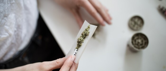 Person die einen Joint dreht vor einem weißen Tisch auf dem ein Grinder steht